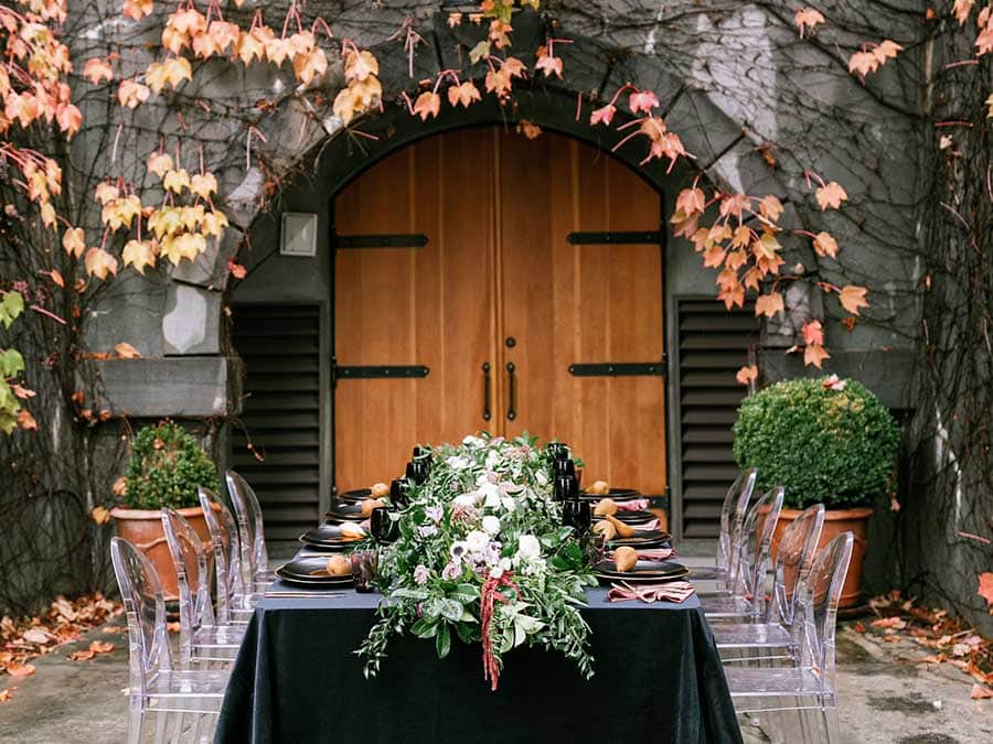 table setting with black table cloth, rose petals, and clear chairs in front of wooden door