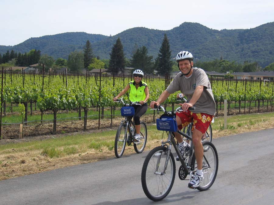 Two people cycle past green vineyards