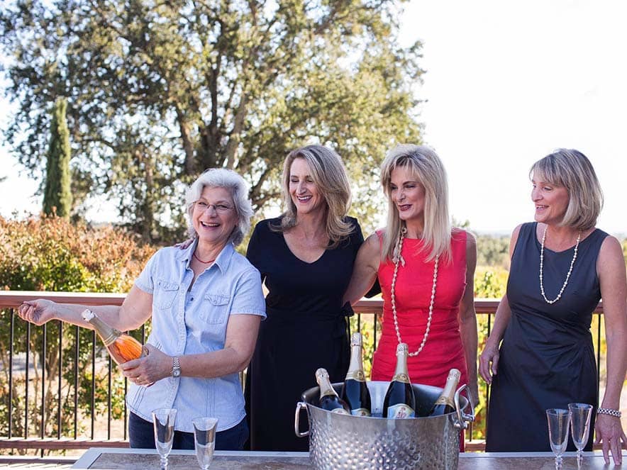 four women standing at a table opening a bottle of wine in celebratin