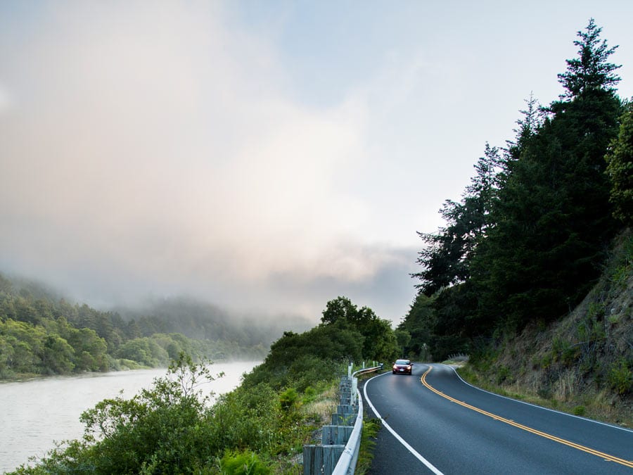 A car drives along a road right next to the river that is covered in fog