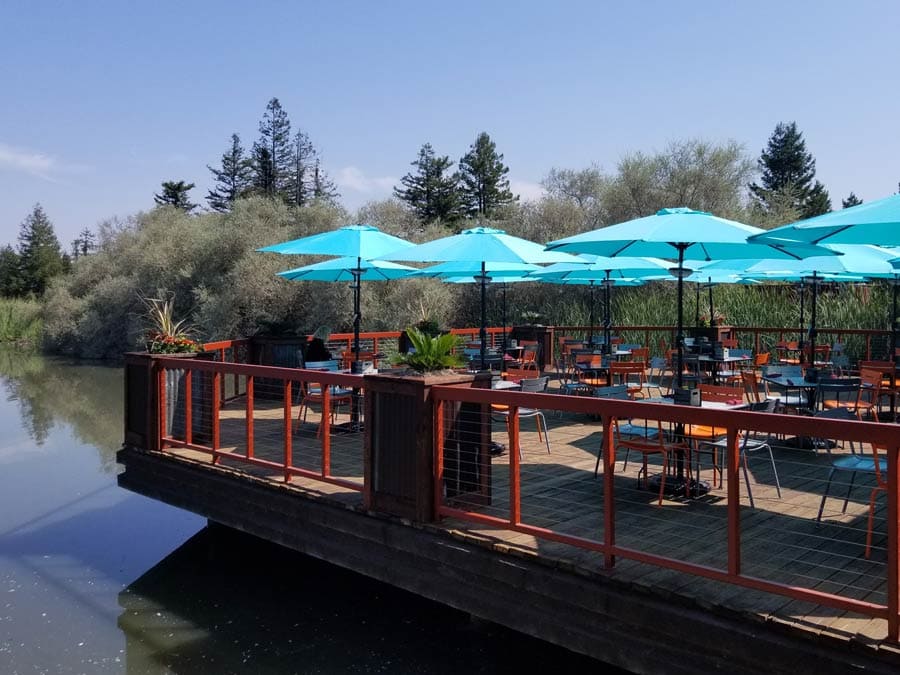 Picnic tables are shaded by blue umbrellas along the lake at the restaurant