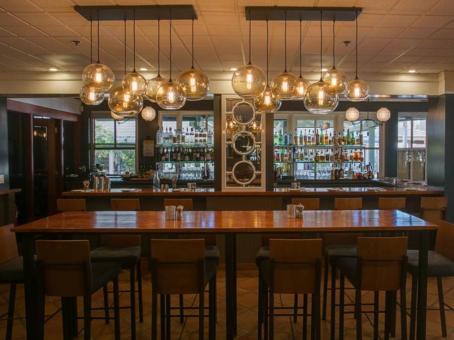 A high top communal table sits in front of the bar under pendant lights