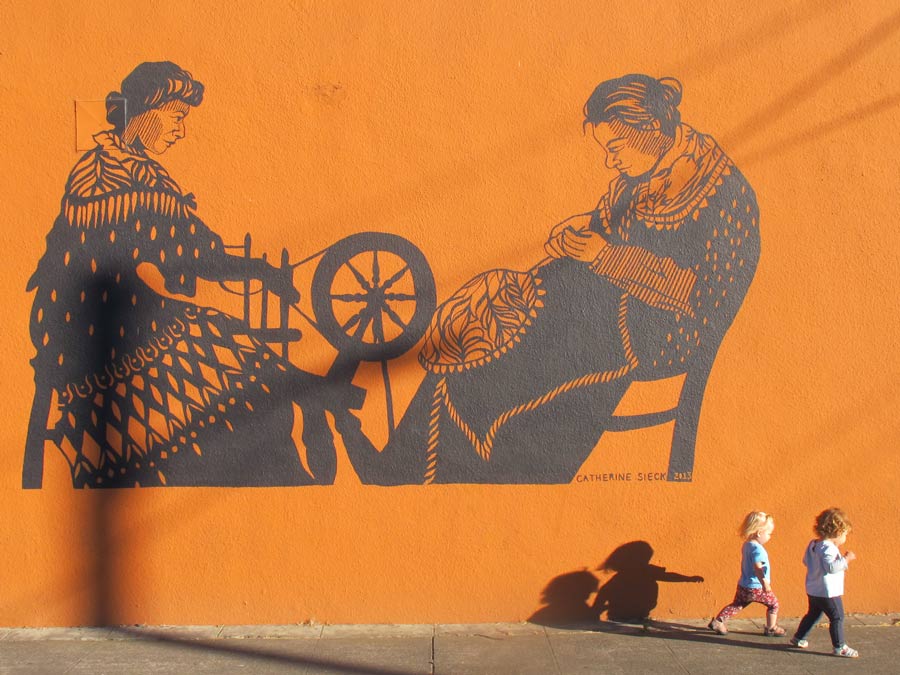 Two children walk by an orange mural of two people spinning wool