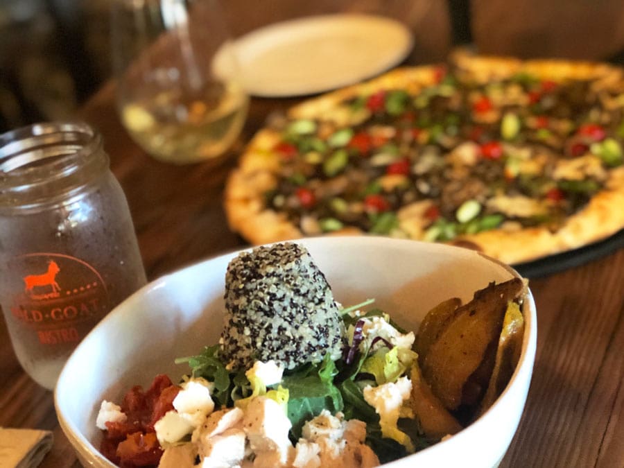 A salad on a table with a pizza in the background