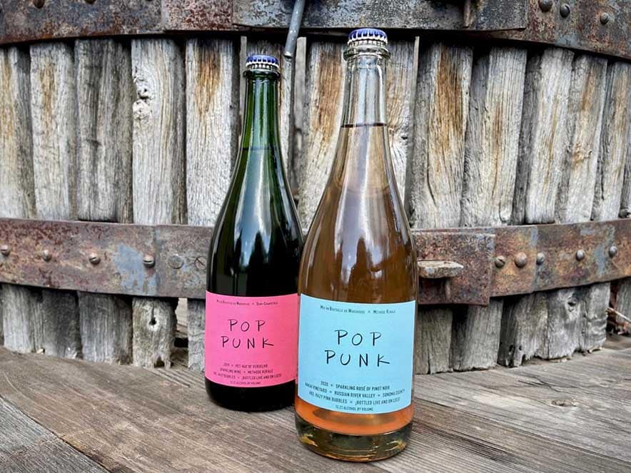 two bottles of pop punk wine with bright labels in front of wine barrel