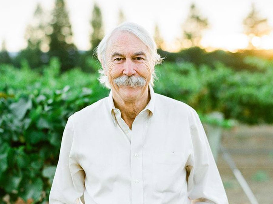 A man with a grey beard and mustache stands in front of green vineyards